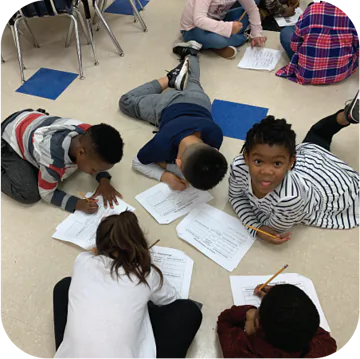 A group of kids are sitting on the floor in a classroom. They are writing and drawing on papers. One girl is making a funny face.