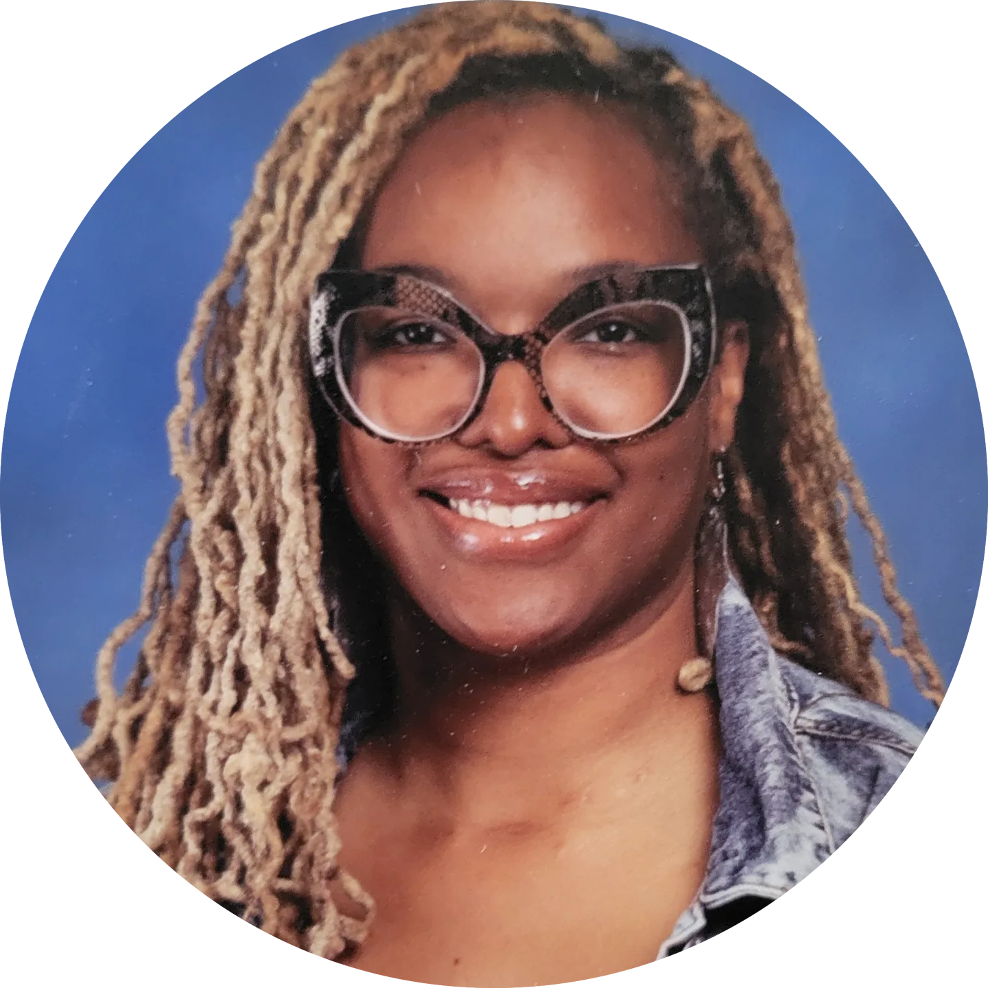 A smiling person with long, braided hair wearing large, round glasses and a denim jacket is centered against a blue background. Their joyful expression radiates warmth and friendliness.