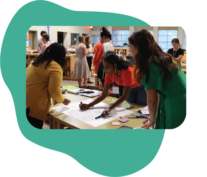 A group of people is engaged in a creative activity around a large table in a bright room. Two women are working closely together; one is drawing on a large sheet of paper while the other assists. There are various colorful shapes and tools spread across the table, suggesting a collaborative project. In the background, more people are interacting and observing. The atmosphere is lively and focused.