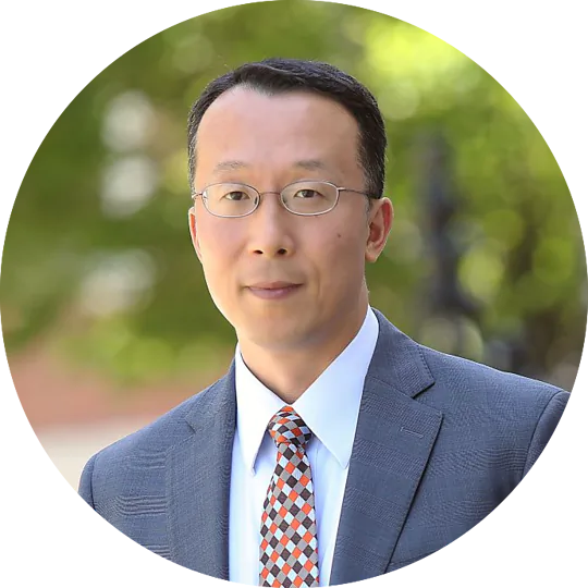 A man with short black hair and glasses stands outdoors, wearing a light blue dress shirt and a gray suit jacket. He has a patterned tie with orange, blue, and white colors. The background is blurred greenery, giving a natural setting.