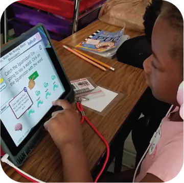 A young girl with headphones is focused on a tablet positioned on a wooden table. The screen displays instructions that read: "Catch the Iguana! I'm going to school! Click on the iguana with the word." Below, there are three green iguana images to choose from. Nearby, there are school supplies like pencils and a worksheet.