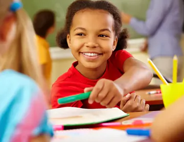 A joyful girl with curly hair smiles while reaching out with a green marker. She is wearing a red shirt and sitting at a classroom desk surrounded by colorful school supplies and books. In the background, a teacher is helping students.