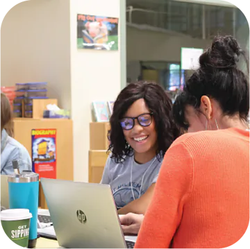 Two students are sitting at a table, smiling and engaged in conversation while working on a laptop. One student is wearing glasses and has curly hair, while the other is wearing a bright orange sweater. In the foreground, there is a green cup with the text "GET SIPPING." In the background, there's a colorful bulletin board with various educational materials.